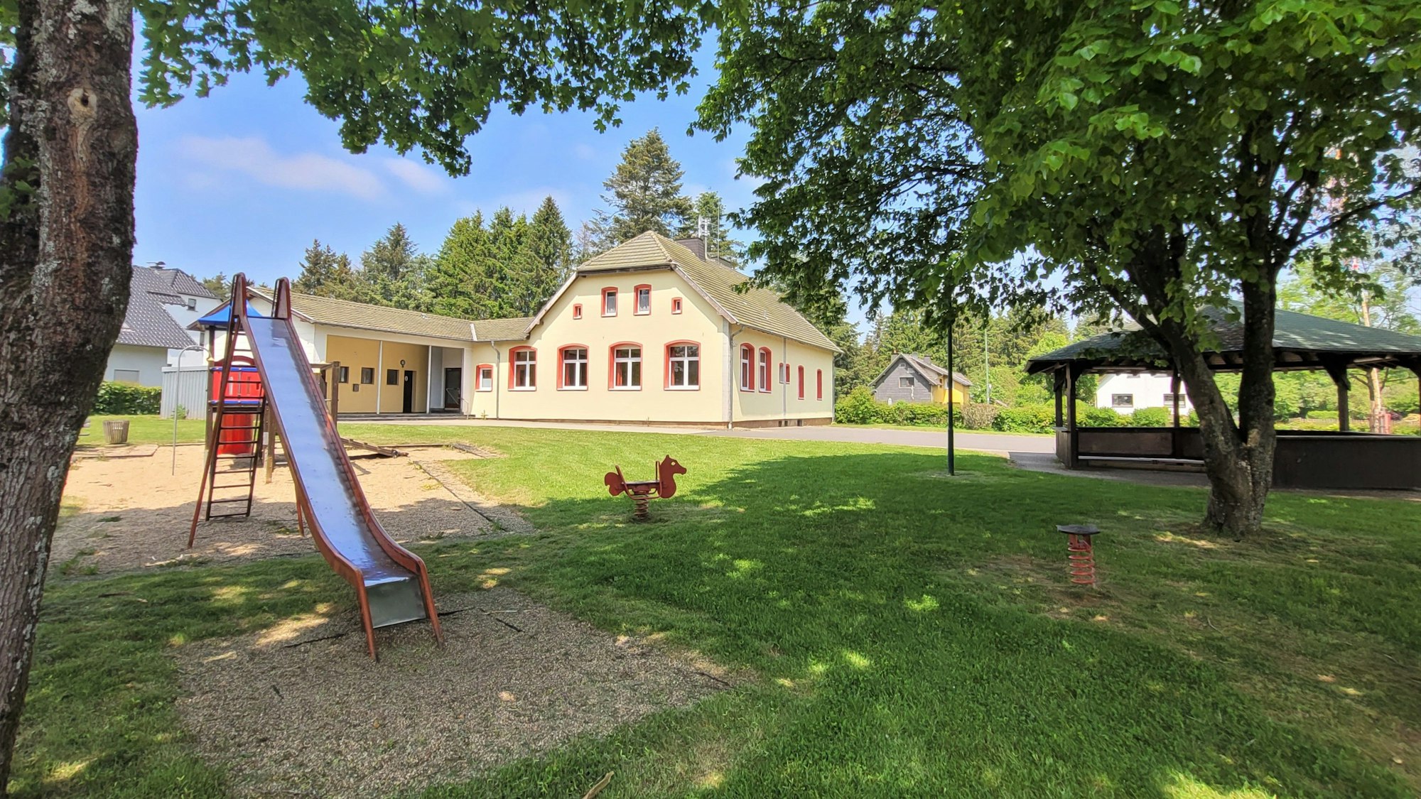 Das Bild zeigt den Dorfplatz in Broich mit einer Rutsche und einem Grillplatz. Im Hintergrund ist das Dorfgemeinschaftshaus zu sehen.