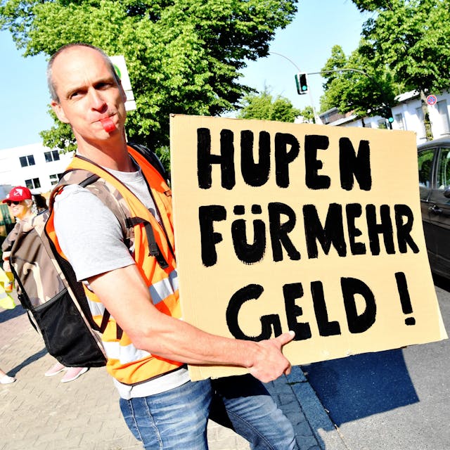 Ein Mitarbeiter einer Berliner Süßwarenfirma steht bei einem Warnstreik vor der Niederlassung von Bahlsen. Er hält ein Schild hoch mit der Aufschrift: „Hupen für mehr Geld!“