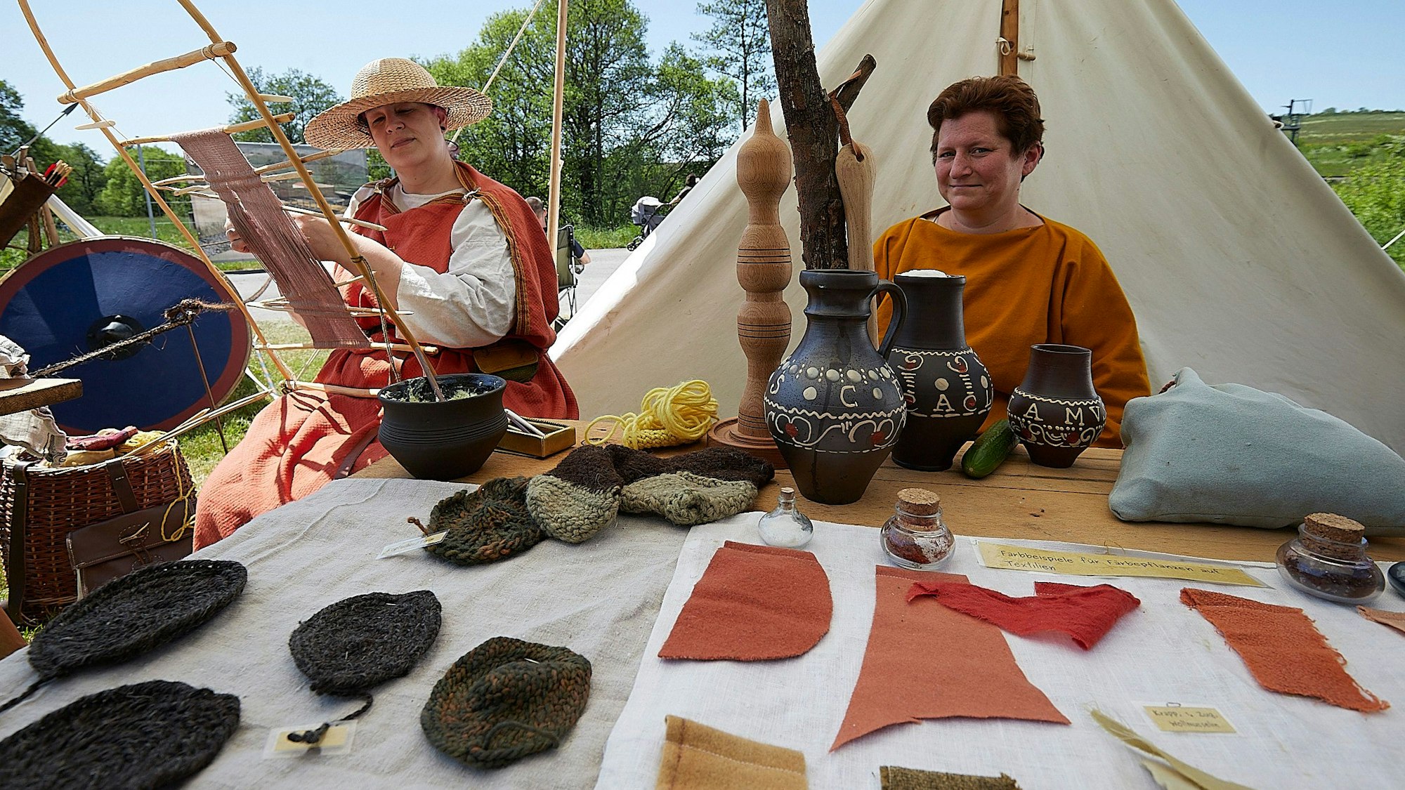 Zwei Frauen sitzen in römischer Kleidung hinter einem Tisch, auf dem sie ihre Tonkrüge und Textil-Produkte ausstellen.