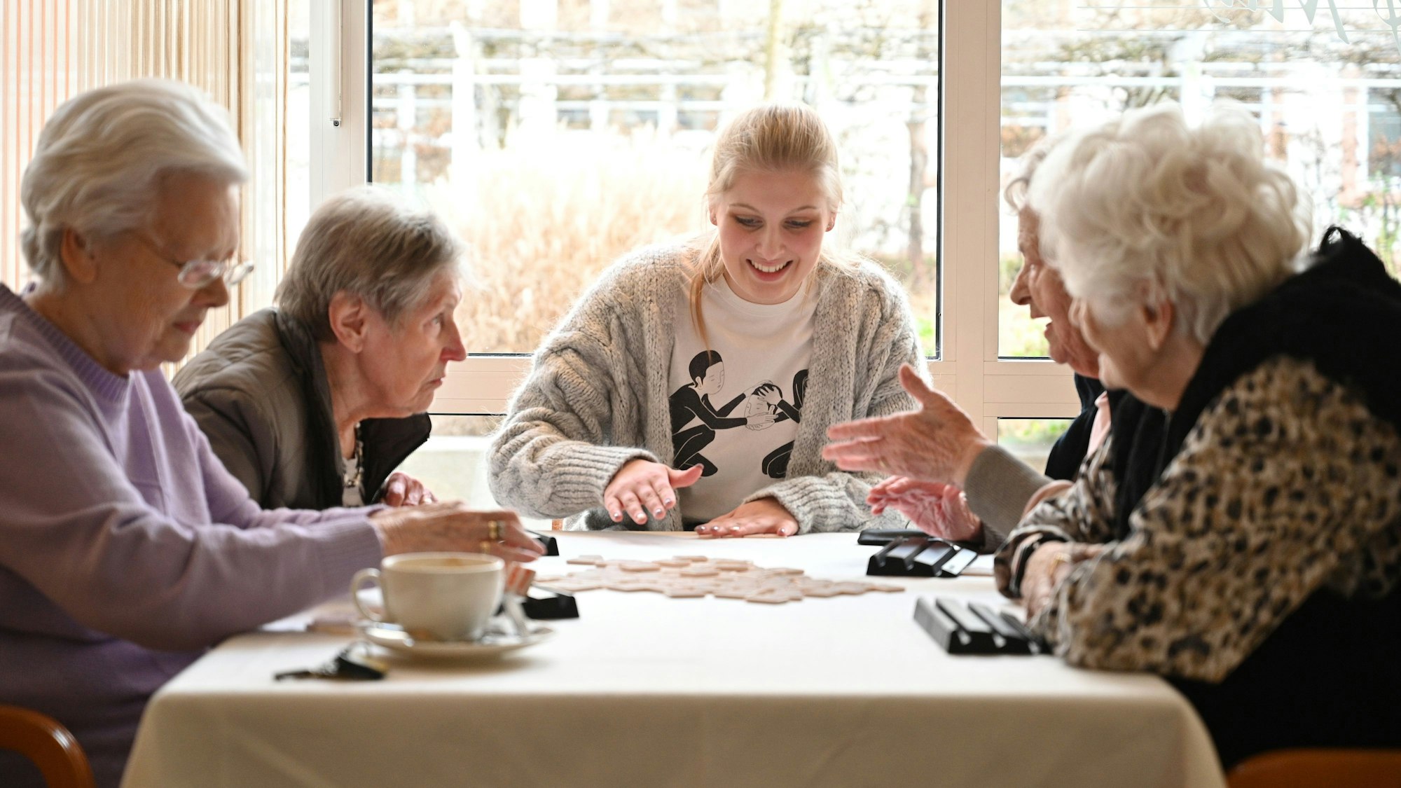 Eine junge Frau sitzt mit vier Bewohnerinnen einer Seniorenresidenz am Tisch und spielt mit ihnen ein Gesellschaftsspiel.