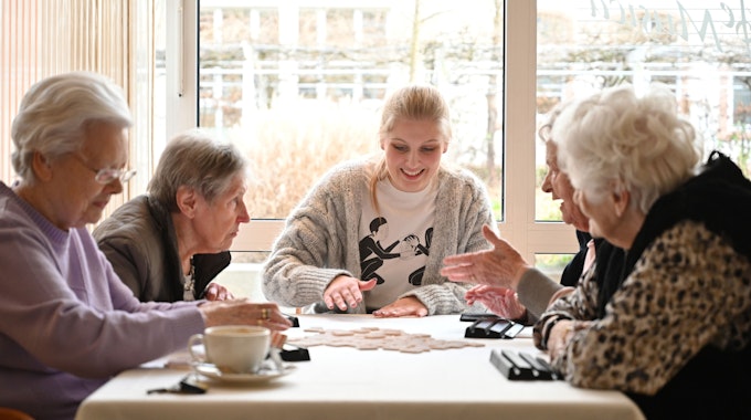 Eine junge Frau sitzt mit vier Bewohnerinnen einer Seniorenresidenz am Tisch und spielt mit ihnen ein Gesellschaftsspiel.&nbsp;