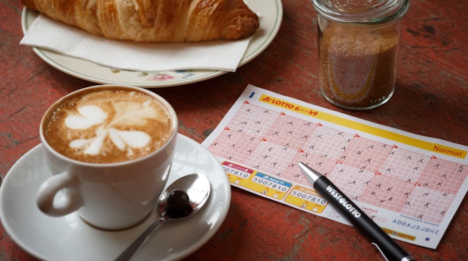Lotto 6aus49 Spielschein auf Tisch neben Kaffeetasse und Croissant.