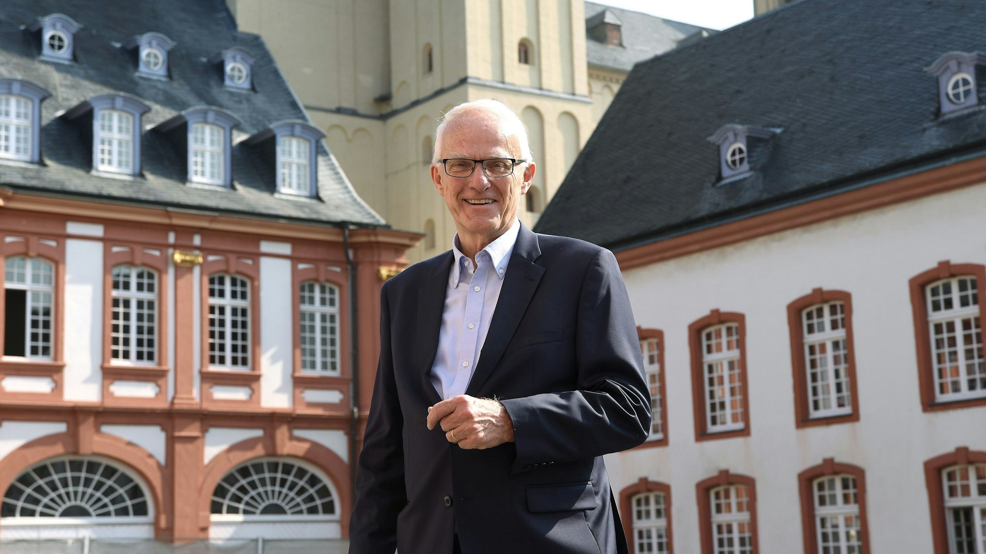 Ministerpräsident a.D. Prof. Dr. Jürgen Rüttgers steht vor mehreren Gebäuden und lächelt in die Kamera.