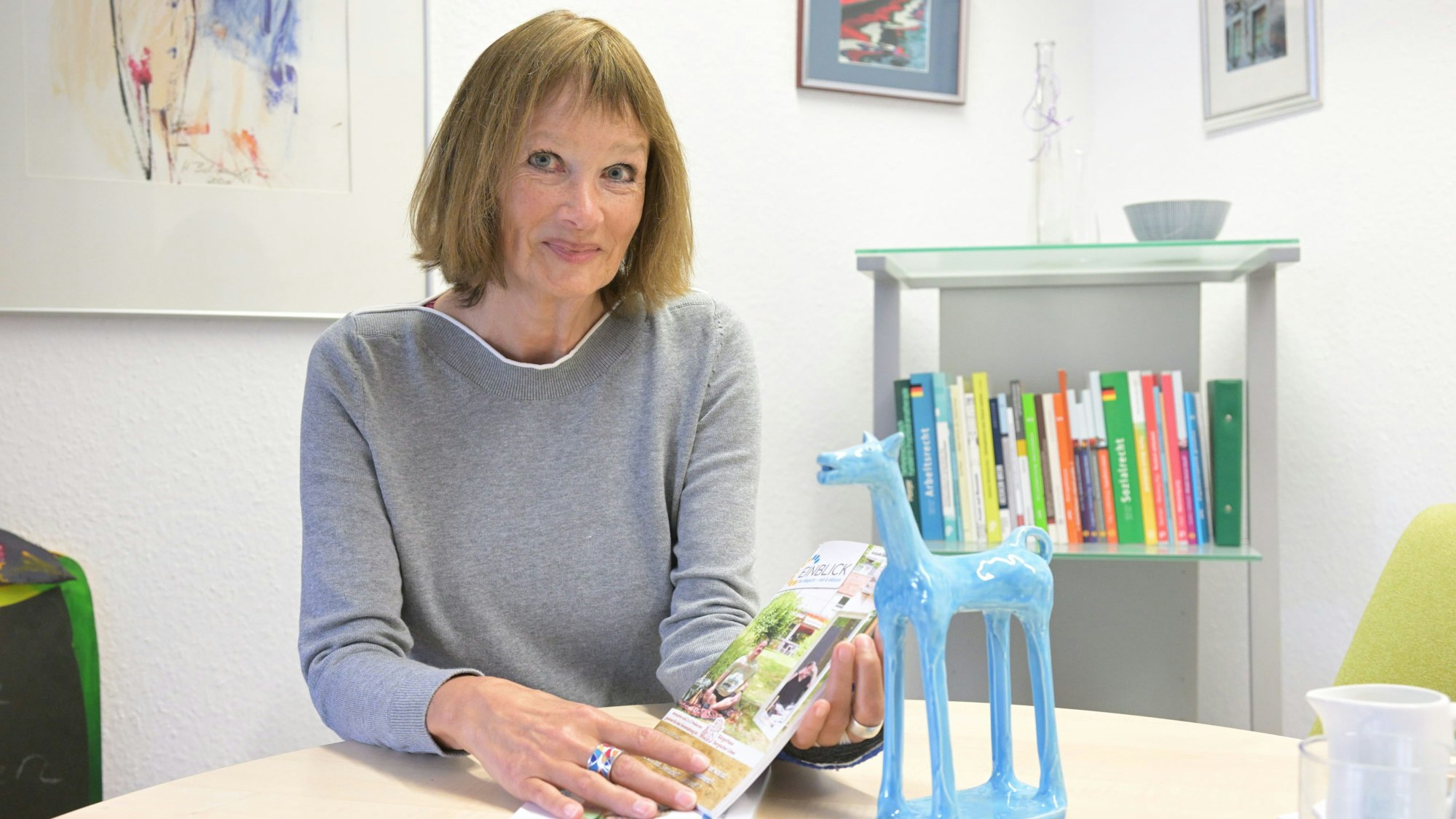 Porträt Claudia Seydholdt, sie sitzt am Tisch und blickt in die Kamera. Neben ihr steht eine blaue Pferde-Figur.