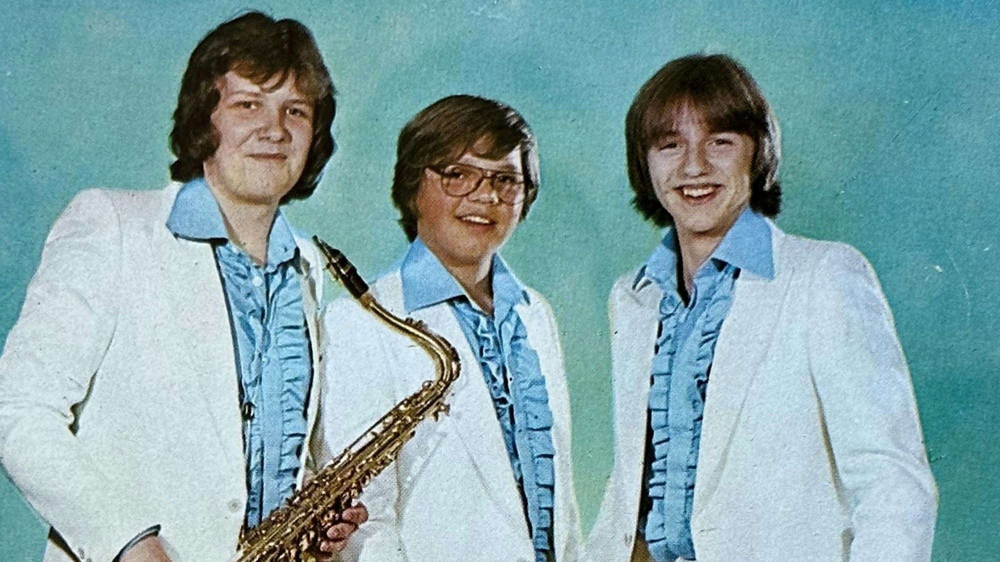 Die drei Bandmitglieder tragen blaue Rüschenhemden und weiße Anzüge.