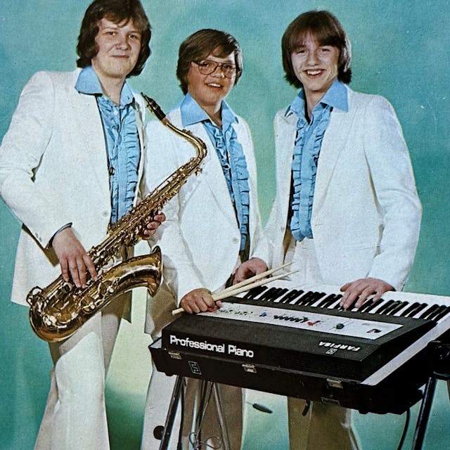 Die drei Bandmitglieder tragen blaue Rüschenhemden und weiße Anzüge.&nbsp;