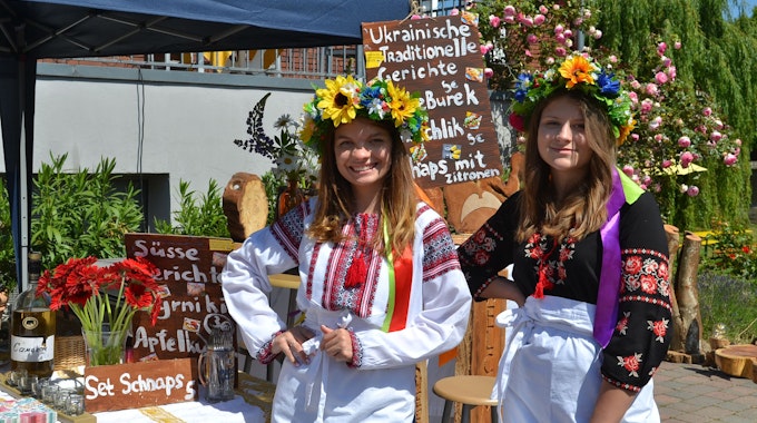 Zwei junge Frauen mit einem bunten Blumenkranz auf dem Kopf stehen vor einem Verkaufsstand auf dem Hoffest des Kulturhofs Velbrück in Metternich.&nbsp;