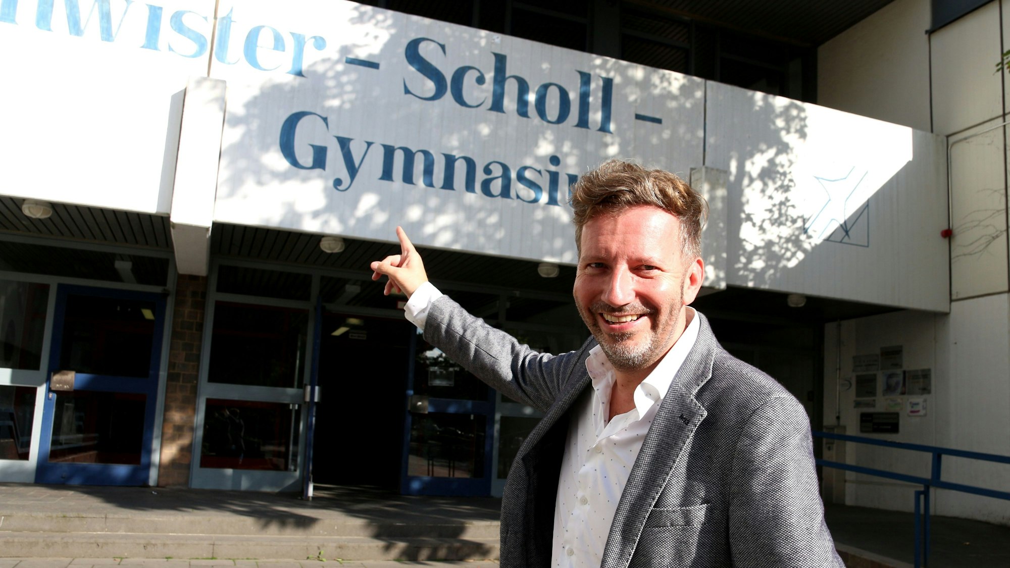 Thorsten Schorn zeigt mit dem rechten Zeigefinger auf das Schild des Geschwister-Scholl-Gymnasiums in Pulheim und schaut dabei lachend in die Kamera.