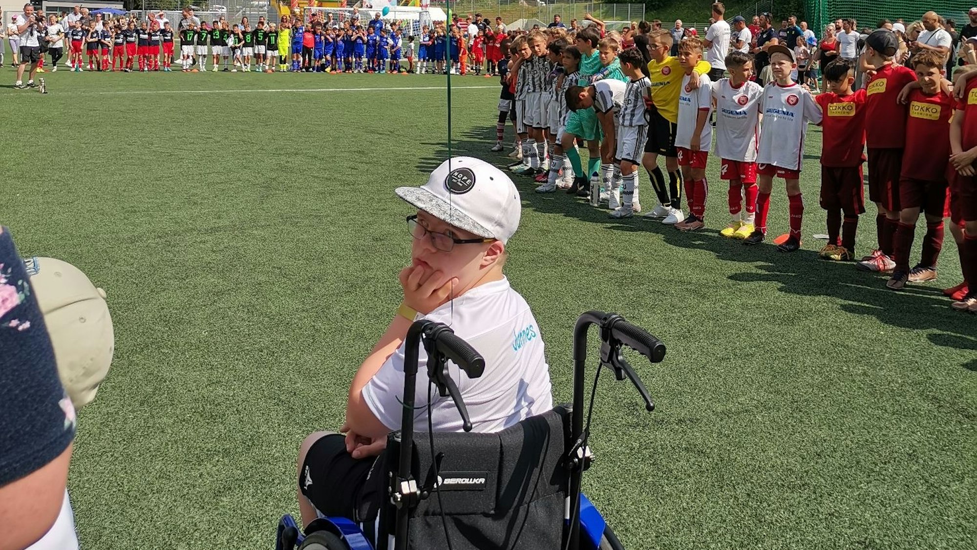 Ein Junge sitzt auf einem Fußballplatz in einem Rollstuhl. Im Hintergrund sind viele weitere Jungs in Trikots zu sehen.