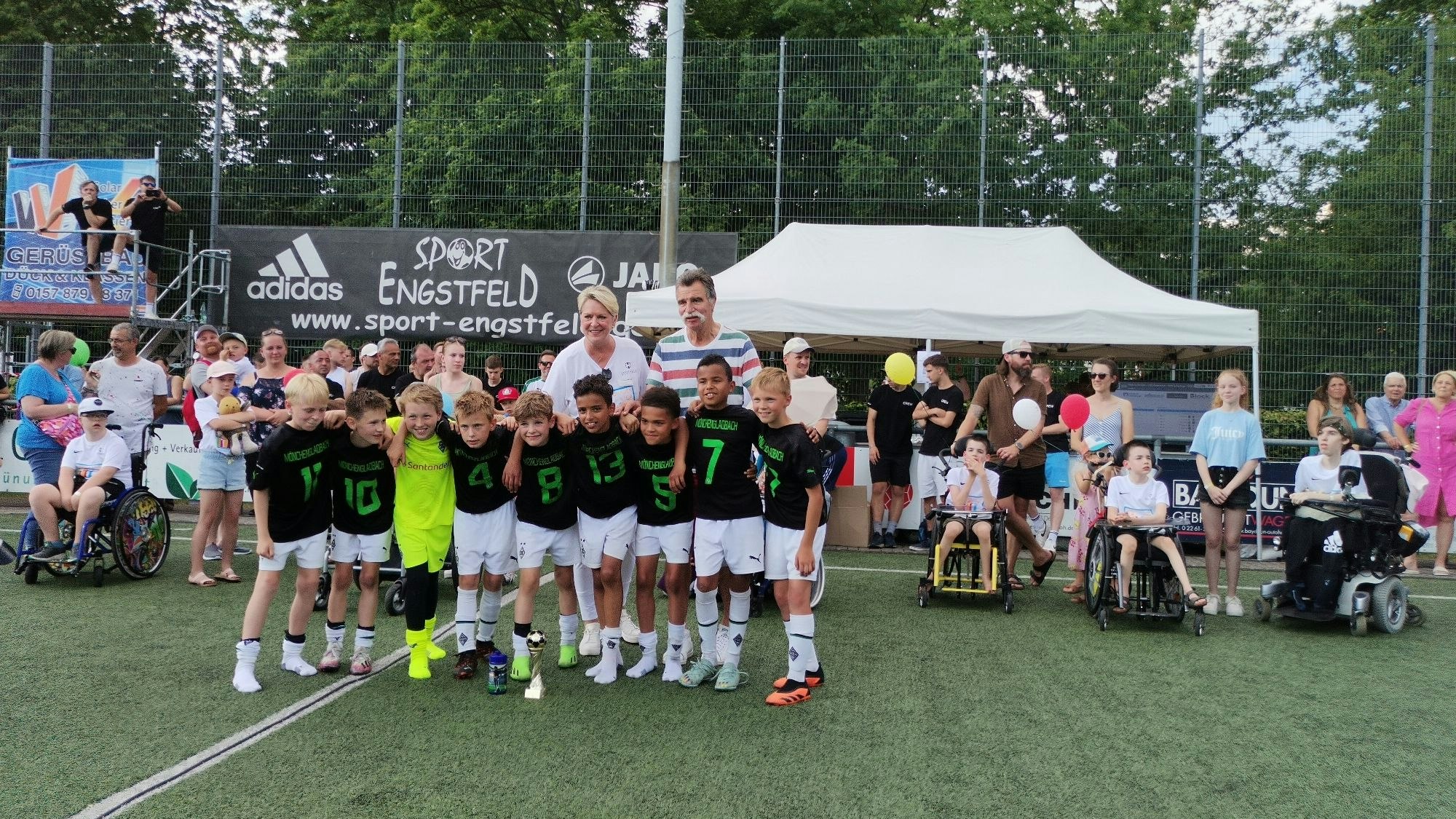 Eine Fußballmannschaft, bestehend aus Jungs, stehen in einer Reihe. Im Hintergrund ist Handballlegende Heiner Brand zu sehen.