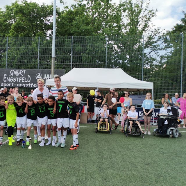 Eine Fußballmannschaft, bestehend aus Jungs, stehen in einer Reihe. Im Hintergrund ist Handballlegende Heiner Brand zu sehen.