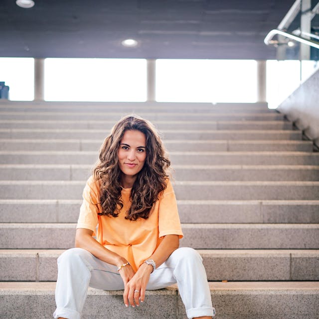 Mona Ameziane sitzt auf einer sterinernen Treppe. Sie hat die Arme auf die Oberschenkel gelehnt und trägt ein oranges Top.