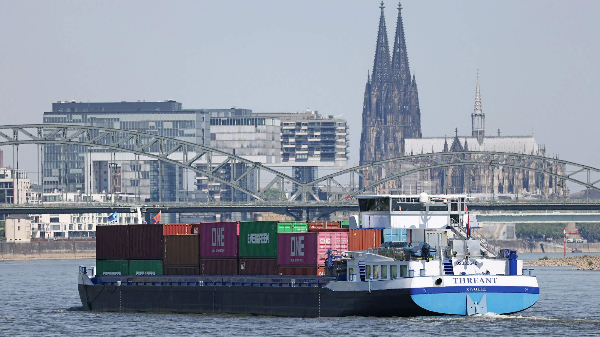 Ein Binnenschiff, mit Containern beladen, fährt in Köln vor dem Dom und den Kranhäusern über den Rhein.