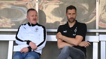 Team-Manager Christofer Heimeroth (r.), hier am 1. Juli 2022 mit Fohlen-Manager Roland Virkus (l.) zu sehen, steht vor seinem Abschied bei Fußball-Bundesligist Borussia Moenchengladbach. Die beiden sitzen auf der Ersatzbank und schauen Richtung Spielfeld.