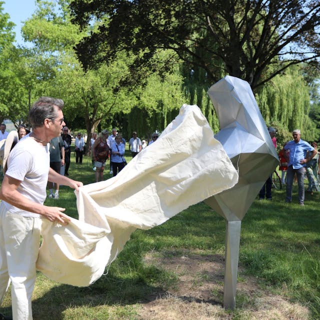 Ein Mann enthüllt ein etwa zwei Meter großes, silbernes Kunstwerk.Im Hintergrund stehende Menschen schauen zu.