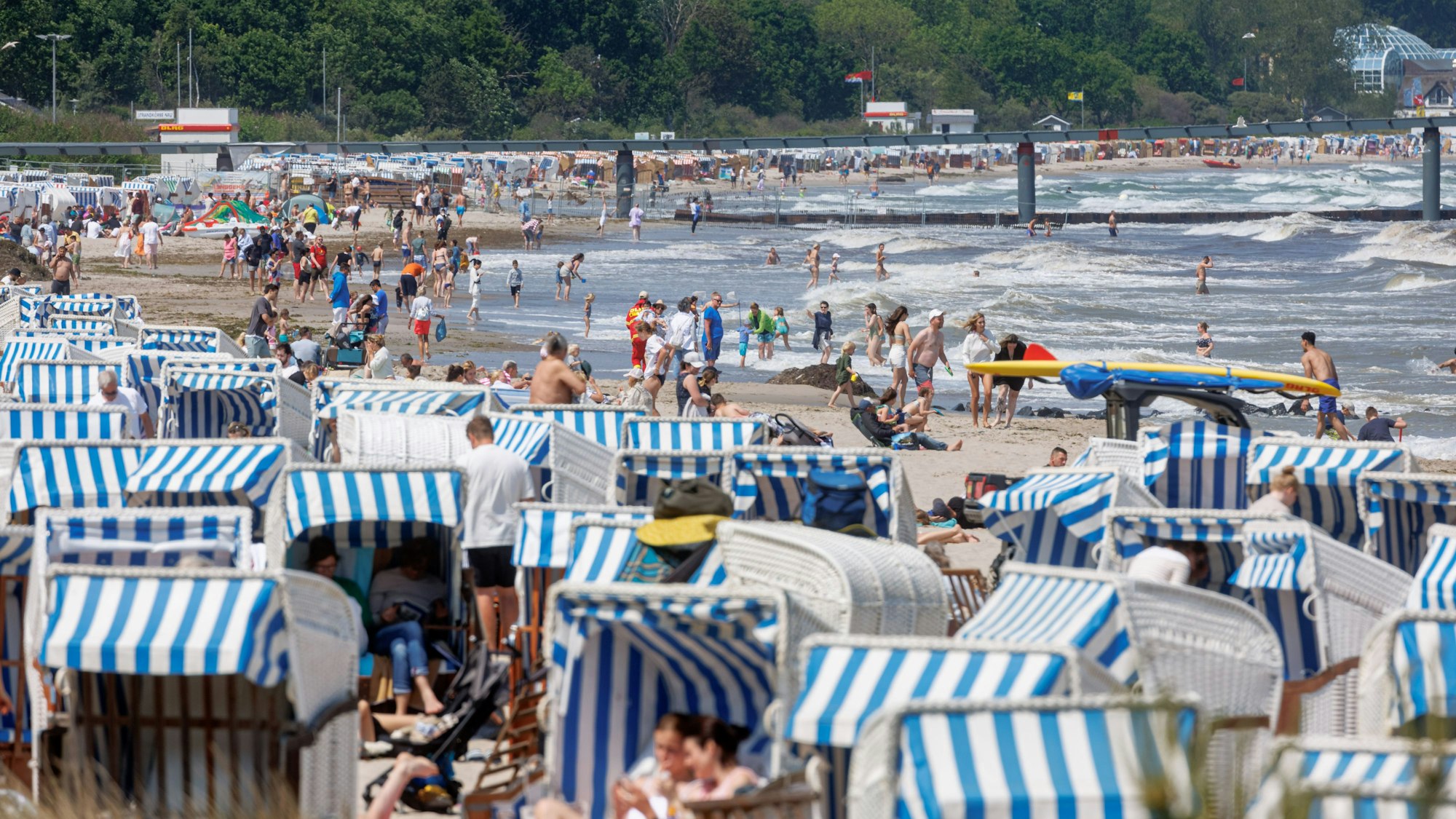 Viele Menschen sind am Strand unterwegs. Einige sitzen in blau-weißen Strandkörben.