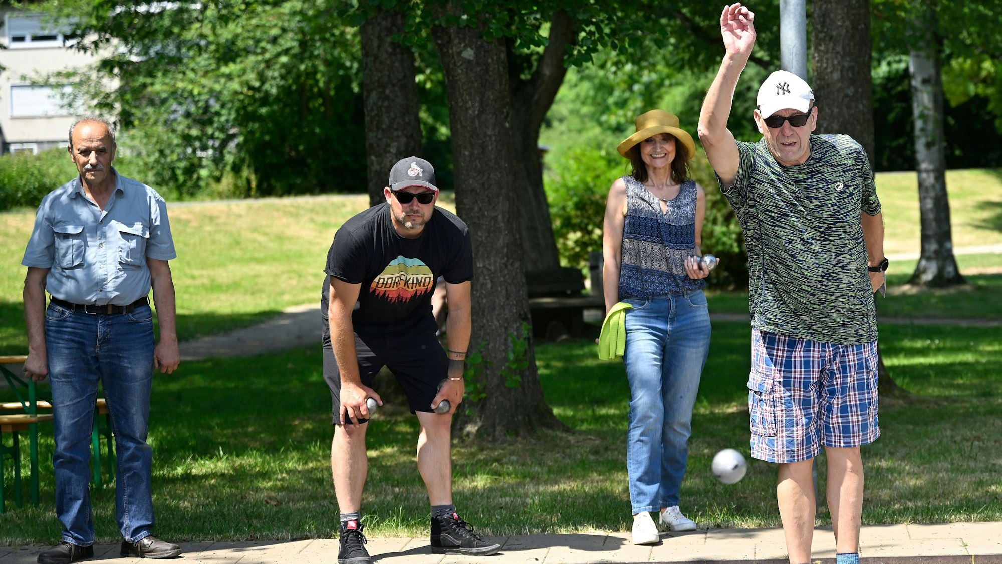 Zwei Männer mit Sonnenbrillen werfen Kugeln in einem Park, im Hintergrund steht eine Frau mit einem Hut und ein Mann.