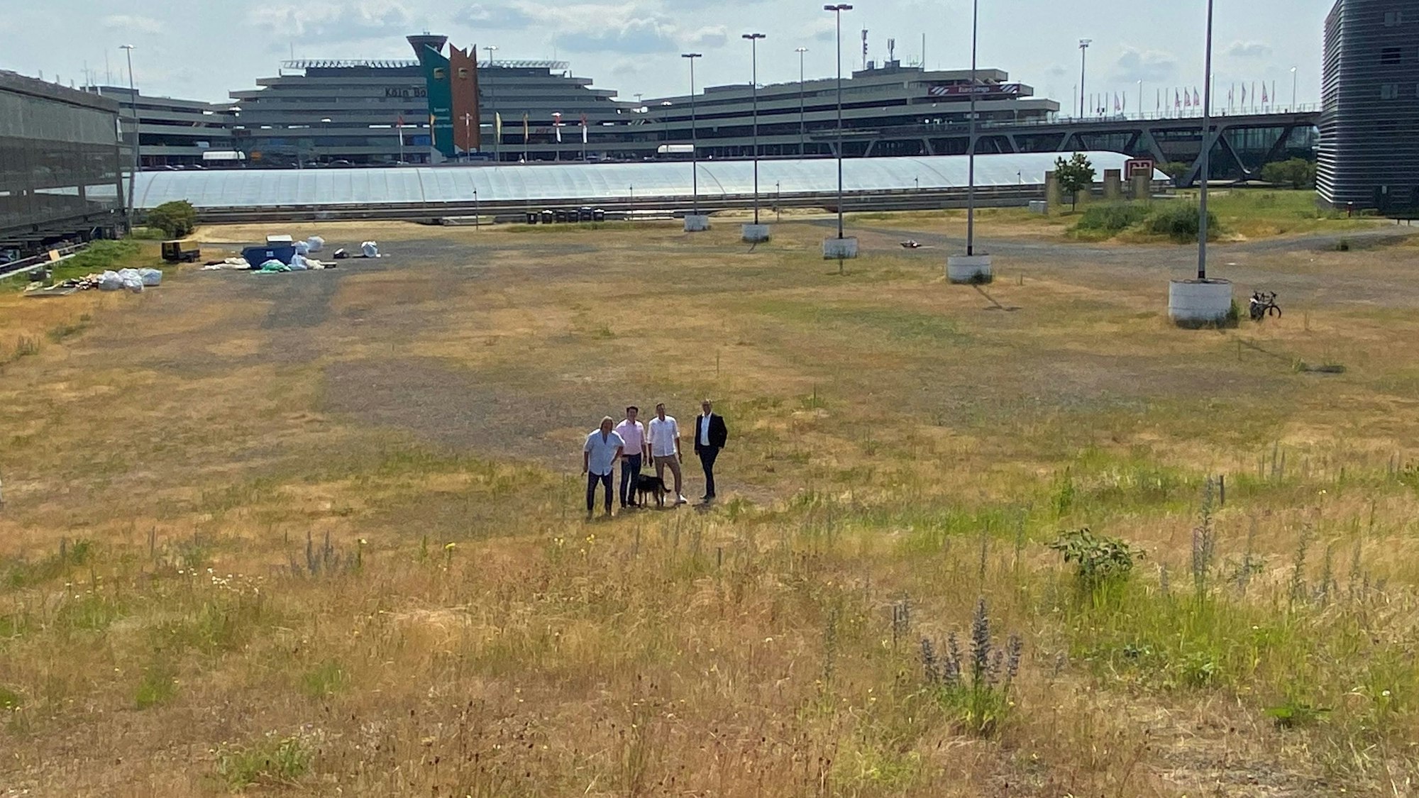 Vier Menschen stehen auf einer großen leeren Fläche, im Hintergrund sind die Flughafen-Gebäude zu sehen.