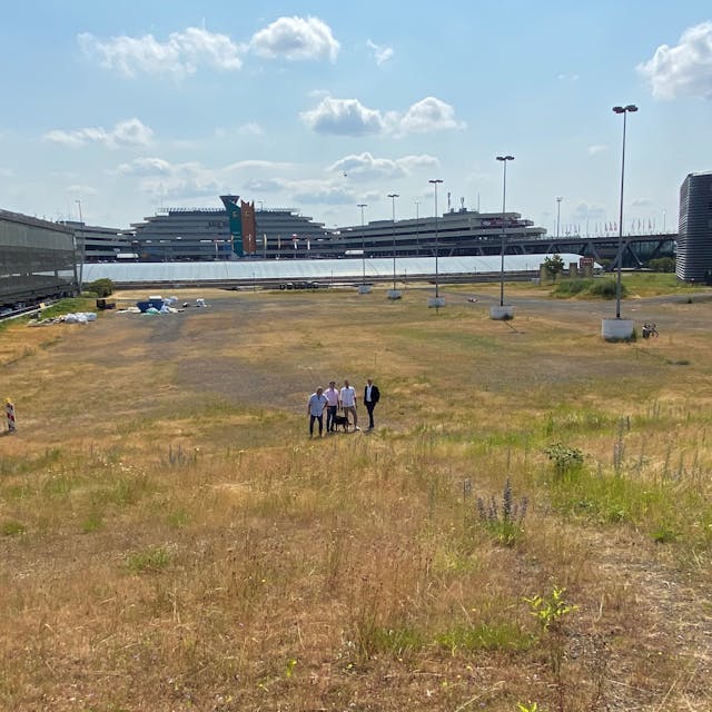 Vier Menschen stehen auf einer großen leeren Fläche, im Hintergrund sind die Flughafen-Gebäude zu sehen.