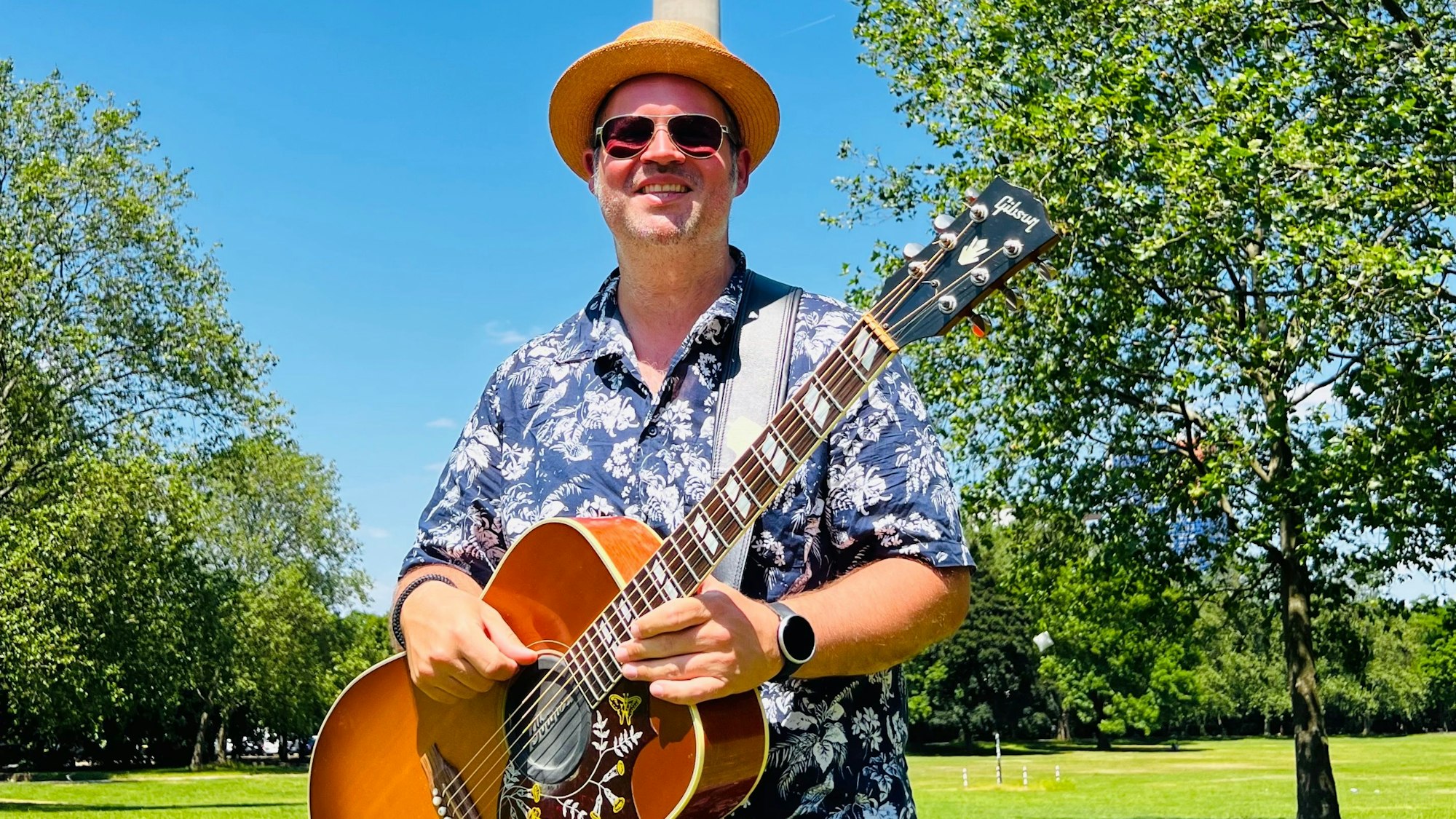 Björn Heuser steht im Hawaiihemd mit Gitarre im Kölner Grüngürtel.