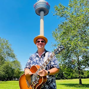 Björn Heuser steht im Hawaiihemd mit Gitarre im Kölner Grüngürtel.