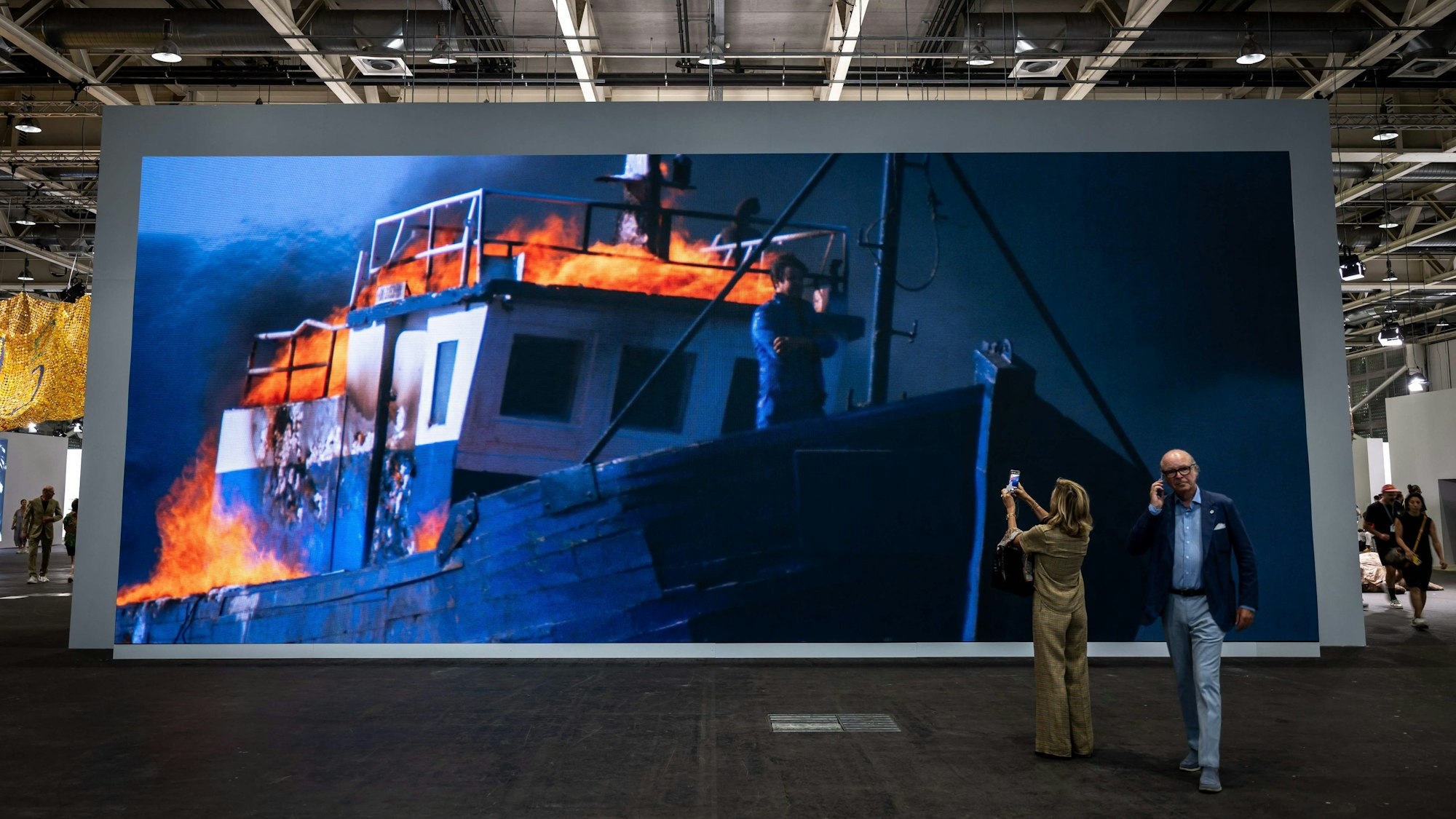 Besucher fotografieren das große Bild. Darauf ist der Künstler am Bug eines brennenden Schiffs zu sehen.