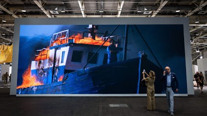 Besucher fotografieren das große Bild. Darauf ist der Künstler am Bug eines brennenden Schiffs zu sehen.&nbsp;