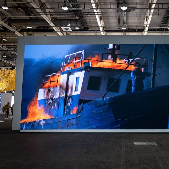 Besucher fotografieren das große Bild. Darauf ist der Künstler am Bug eines brennenden Schiffs zu sehen.&nbsp;
