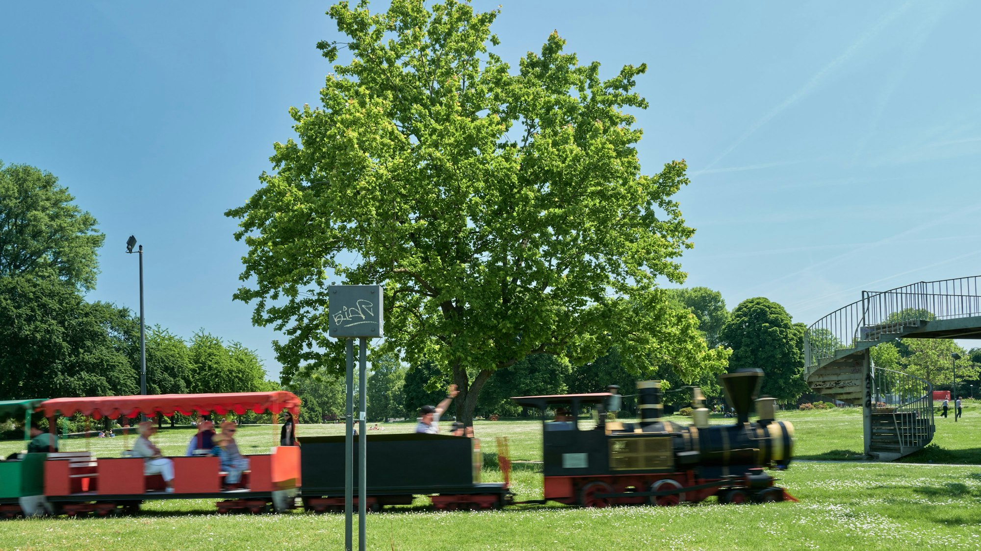 Blick auf den Rheinpark in Köln, wo regelmäßig die Bimmelbahn fährt. Menschen genießen das Grün in Deutz.