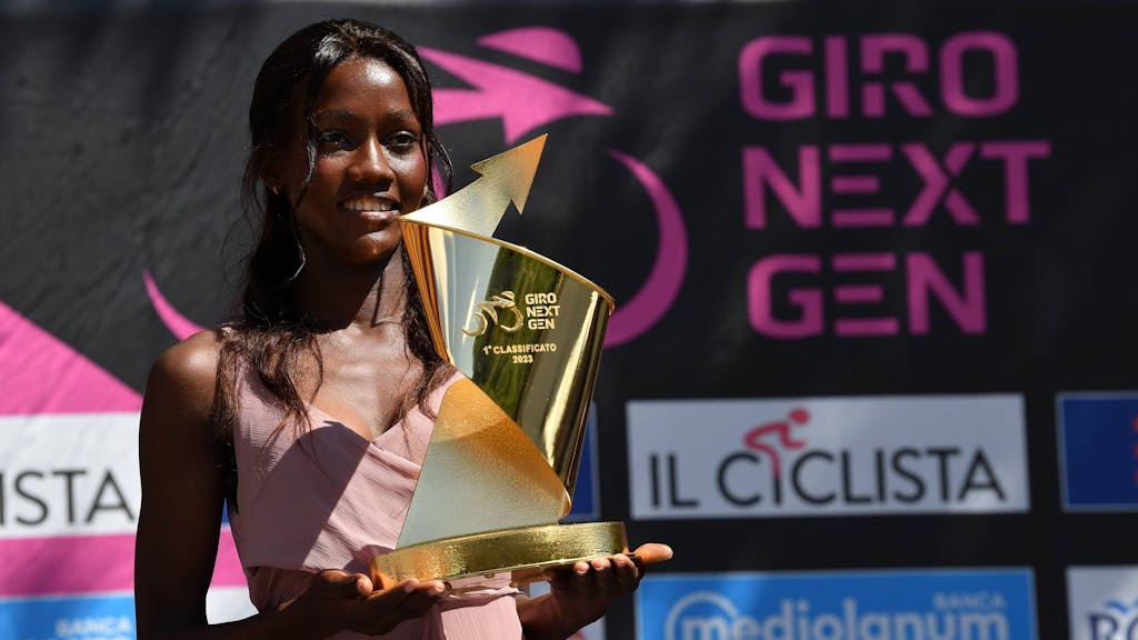 Eine Frau hält einen goldenen Pokal in den Händen. Im Hintergrund ist der Schriftzug Giro Next Gen zu sehen.
