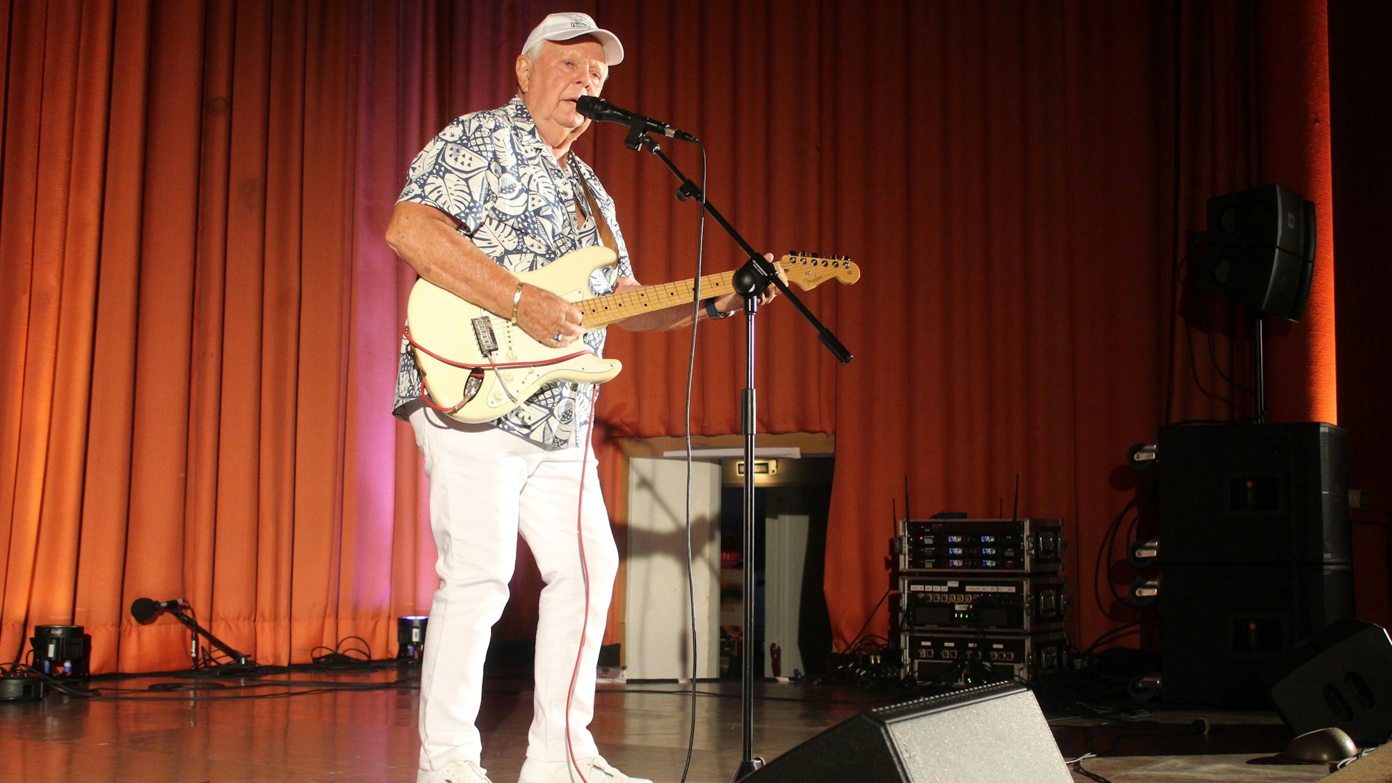 Schlagerstar Graham Bonney steht mit Gitarre auf einer Bühne und singt in ein Standmikro.