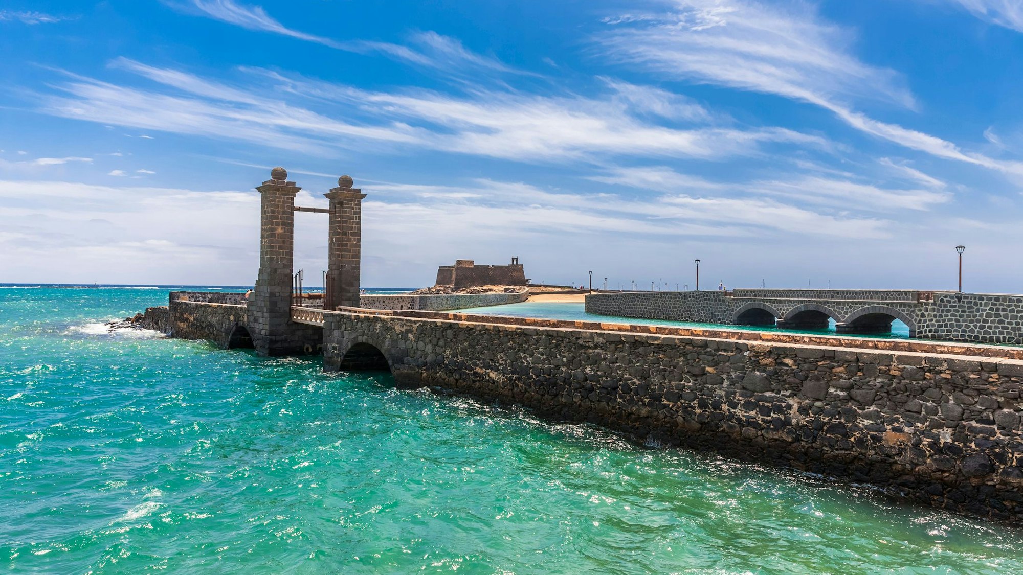 Aus türkisfarbenem Wasser ragt eine Steinbrücke. Im Hintergrund ist eine Festung aus Stein zu sehen.