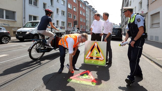 Ein KVB-Mitarbeiter besprüht die Straße mit einem Warnsymbol. Im Hintergrund halten Polizeipräsident Falk Schnabel und Verkehrsdezernent Ascan Egerer das Piktogramm hoch.&nbsp;