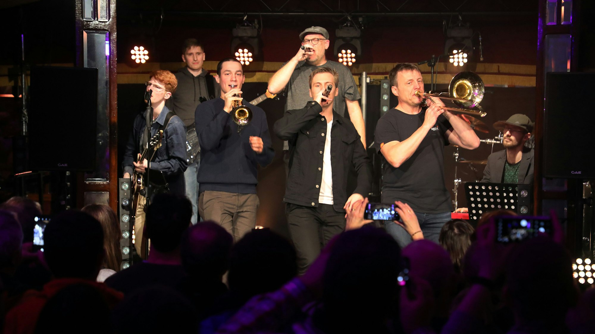 Sieben Männer befinden sich auf einer Bühne, singen und musizieren vor Publikum.