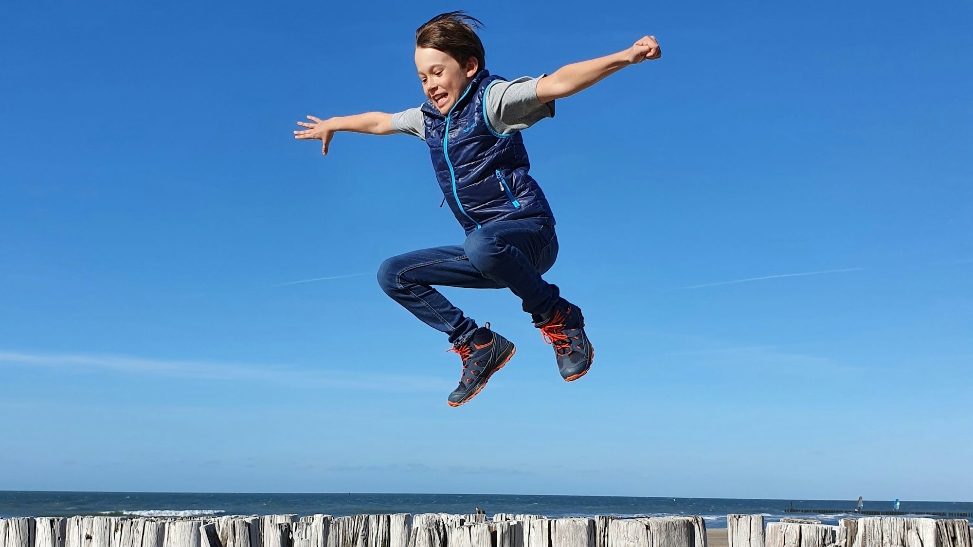 Ein Junge springt über Hölzer an einem Strand vor blauem Himmel und Meereshorizont.