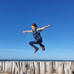 Ein Junge springt über Hölzer an einem Strand vor blauem Himmel und Meereshorizont.
