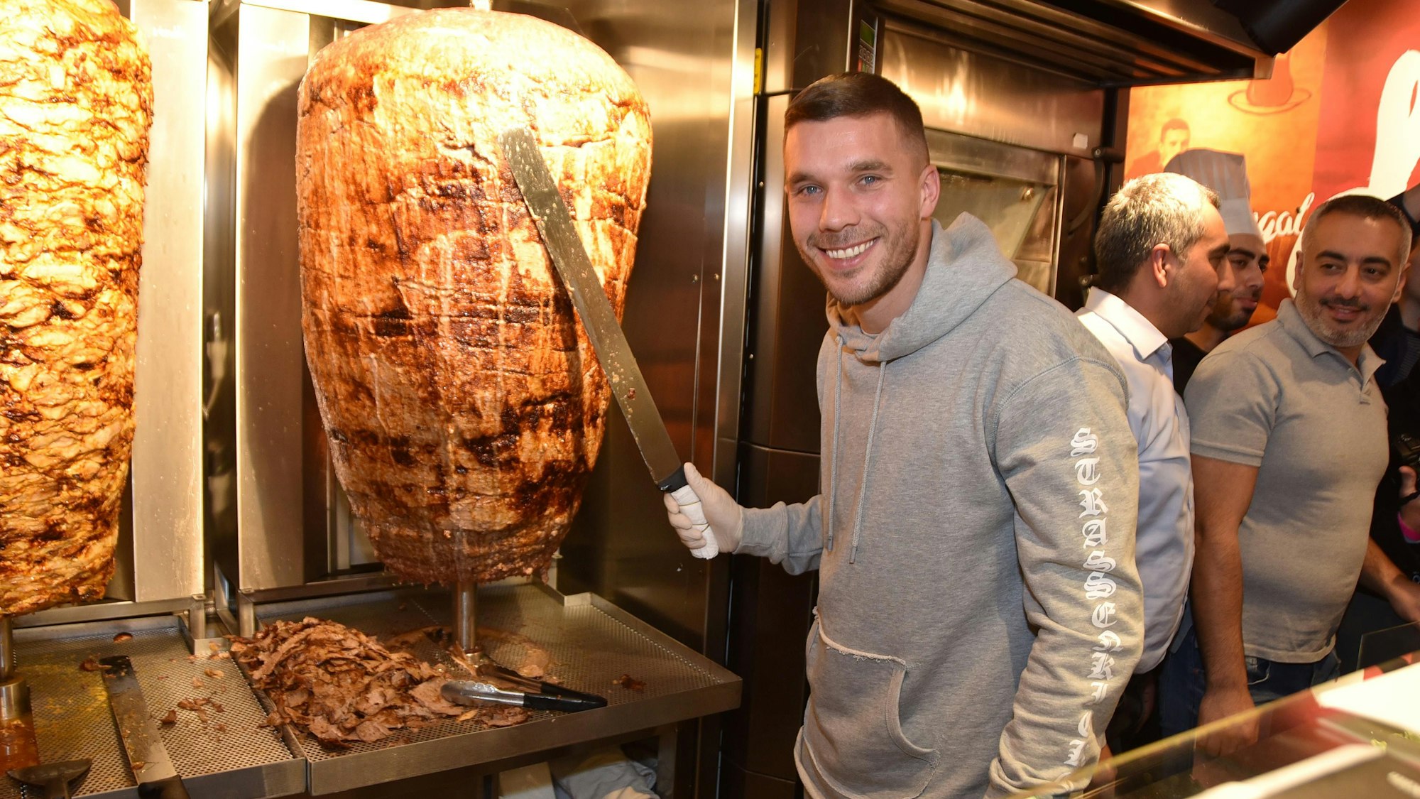 Fußball-Weltmeister Lukas Podolski steht 2018 in Köln bei der Eröffnung eines Döner-Ladens an einem Dönerspieß. In der rechten Hand hält er ein Dönermesser, er schaut in die Kamera.