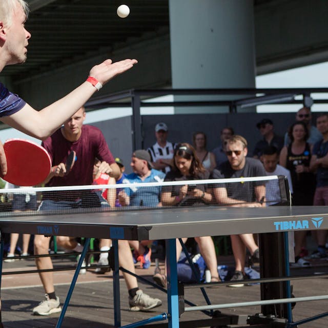 Zwei Tischtennisspieler sind mitten in einem Ballwechsel, die Platte steht draußen auf Holzboden, im Hintergrund sind Zuschauer zu sehen.