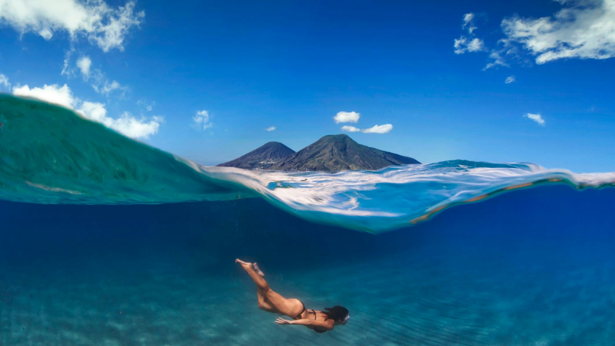 Eine Frau taucht im Meer, über ihr ist eine Insel mit zwei Vulkanen zu sehen.