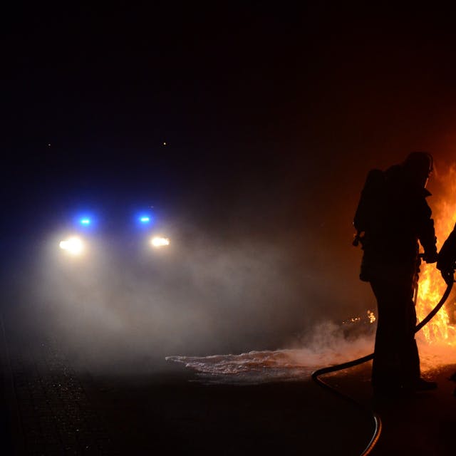 Dunkle Umrisse von zwei Feuerwehrleuten. Im Hintergrund sieht man ein brennendes Autowrack. (Archivbild)