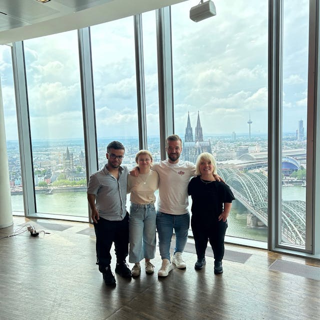 Zu sehen sind vier Menschen. Zwei Frauen und zwei Männer sind zu sehen, die nebeneinander in einem Gebäude mit Glasfassade stehen. Im Hintergrund befindet sich der Kölner Dom.