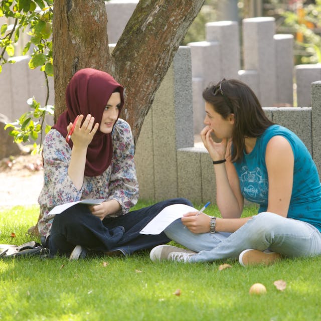 Zwei jugendliche Frauen, eine trägt ein Kopftuch, sitzen im Kölner Garten der Religionen und unterhalten sich.