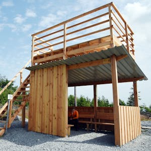 Das Bild zeigt eine Holzhütte, deren Flachdach über eine Treppe bestiegen werden kann.