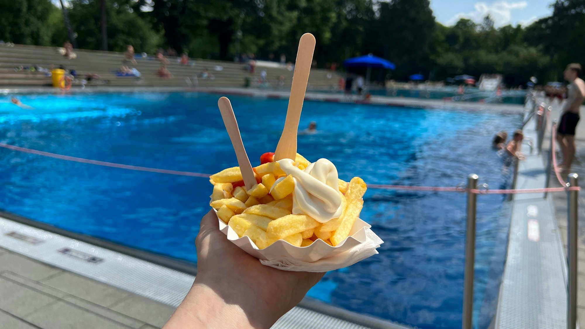 Eine Schale mit Pommes und Ketchup Mayo wird vor das Schwimmbecken im Hintergrund präsentiert.