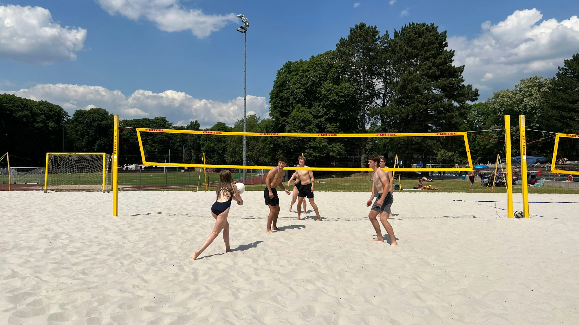 Auf einem Beach-Volleyballfeld spielen vier Männer und zwei Frauen Beach-Volleyball im Freibad.