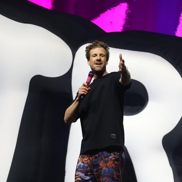 Luke Mockridge steht in einem schwarzen T-Shirt, einer bunten Hose und mit einem pinken Mikrofon in der Kölner Lanxess-Arena auf der Bühne.