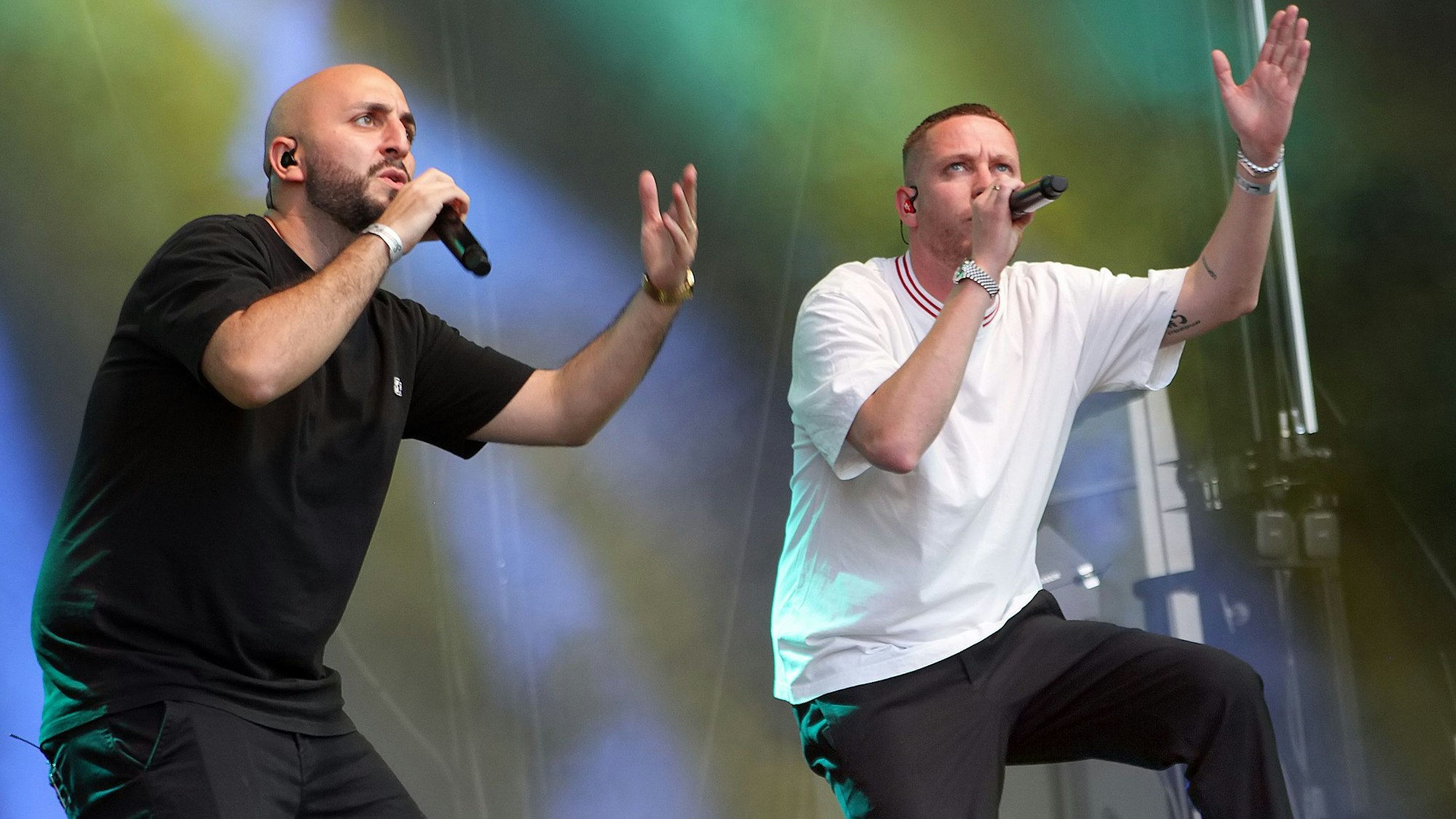 Zwei Männer mit Mikrophonen in der Hand stehen auf einer Bühne. Die linke Hand haben sie jeweils in die Luft gestreckt.
