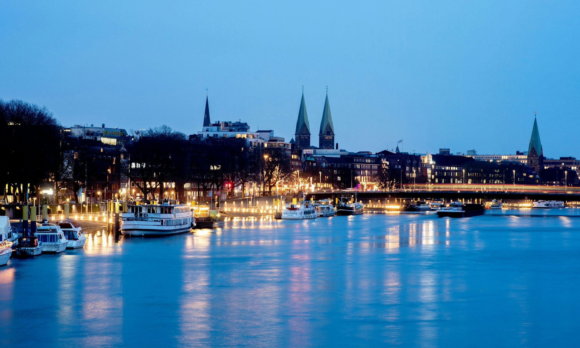 Die Lichter der Uferpromenade spiegeln sich in der Dämmerung in der Weser, während im Hintergrund die Altstadt und der Bremer Dom St. Petri zu erkennen sind.