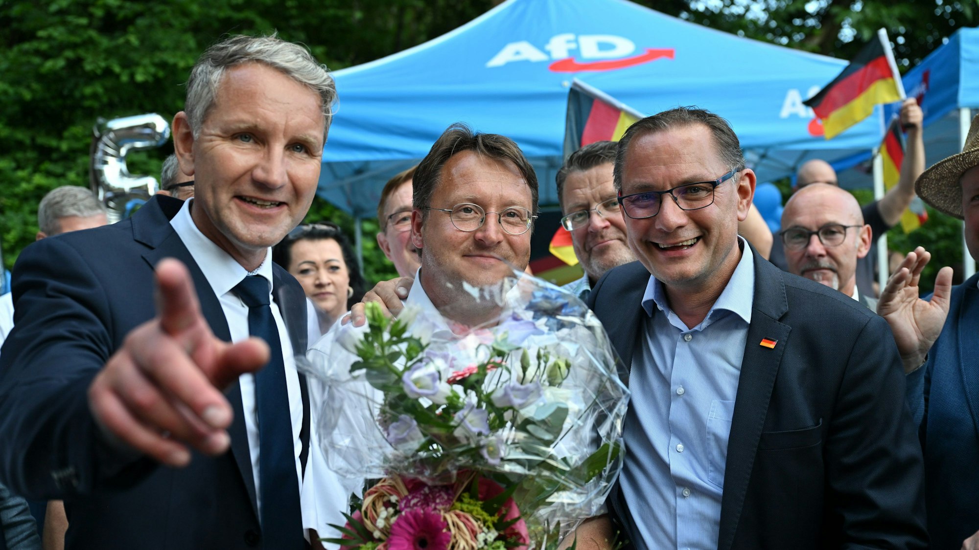 Björn Höcke (links), Vorsitzender der AfD Thüringen, und Tino Chrupalla, AfD-Bundesvorsitzender (rechts), gratulieren dem Wahlsieger Robert Sesselmann. Er hält einen Blumenstrauß in der Hand.