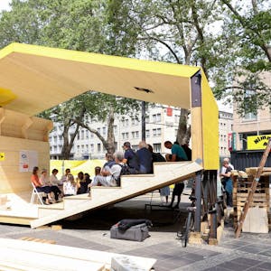 Ein Holzpavillon in gelber Farbe auf einem Platz.